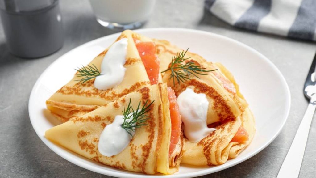 Palacinky preložené do trojuholníkov, plnené lososom, podávané so smotanou a kôprom na bielom tanieri.