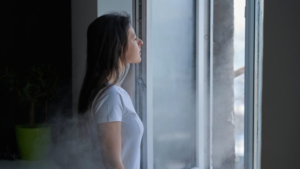 Žena stojí pri otvorenom okne počas chladného dňa a nadýchne sa čerstvého vzduchu.