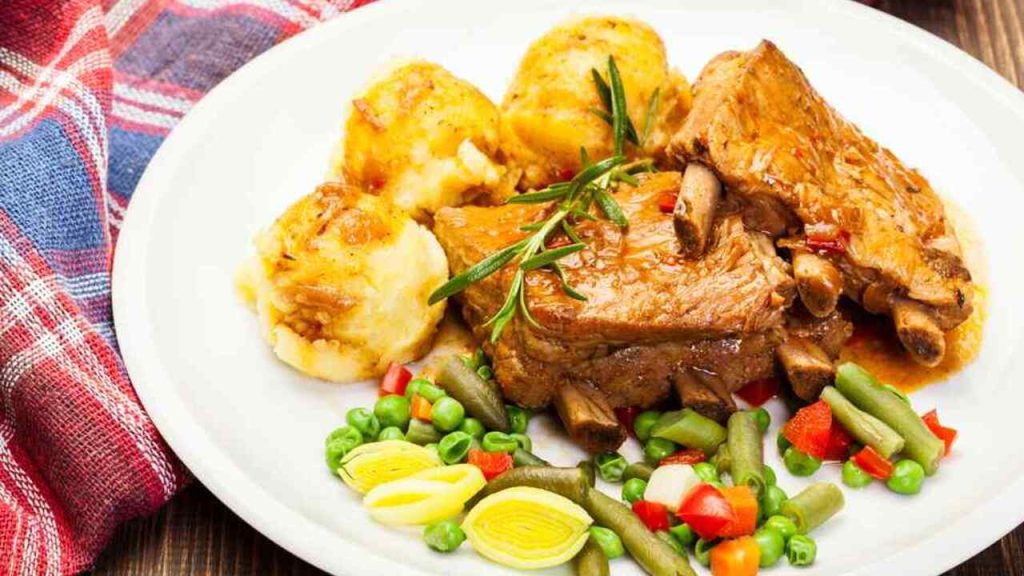 Bravčové rebrá v omáčke servírované so zemiakmi a dusenou zeleninou na bielom tanieri.