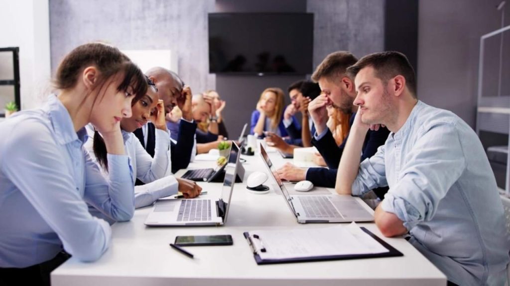 Skupina zamestnancov sedí pri dlhom stole v kancelárii, pôsobia znudene, unavene a bez energie.