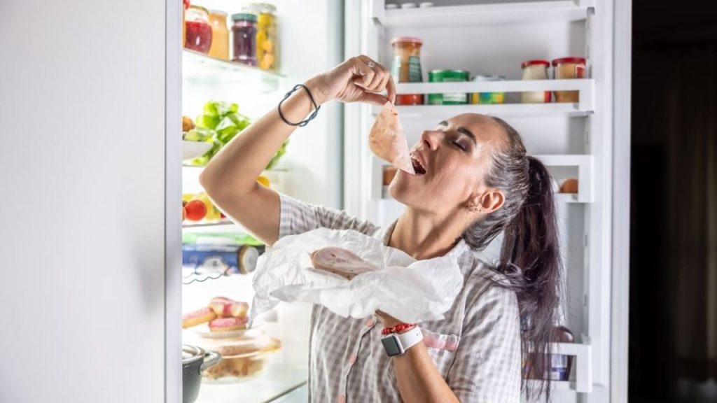 Žena stojaca pri otvorenej chladničke a ochutnávajúca plátok šunky zabalený v papieri.