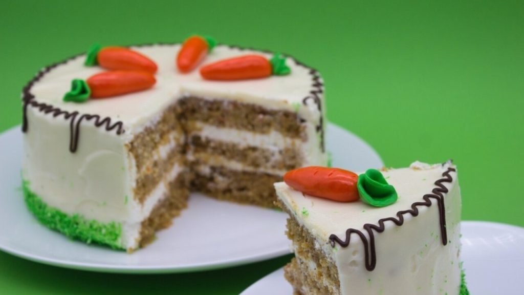 Celý mrkvový tortový koláč s krémom, ozdobený marcipánovými mrkvičkami, s odkrojeným kúskom.