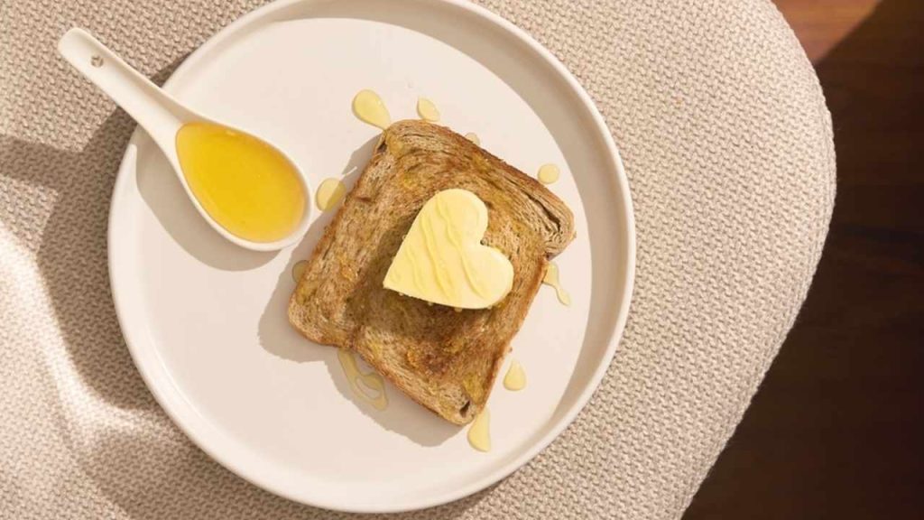 Raňajkový toast s lyžicou medu a maslom v tvare srdca, podávaný ako zdravá voľba pre seniorov.