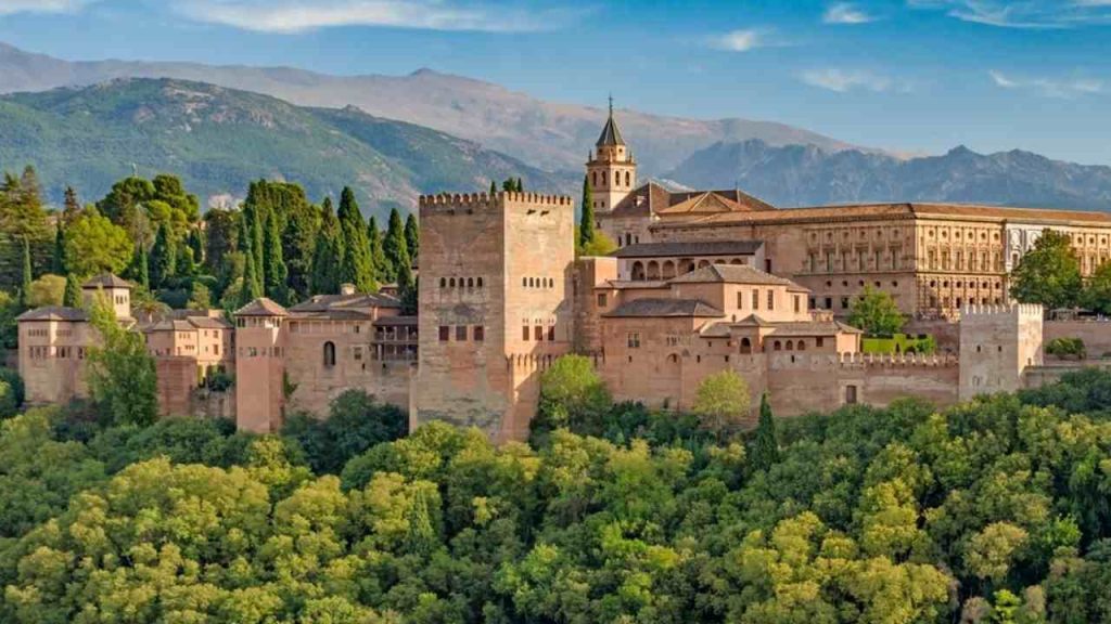 Panoramatický pohľad na pevnosť a palácový komplex Alhambra v Granade, obklopený zeleňou a horami Sierra Nevada počas zlatého svetla západu slnka.
