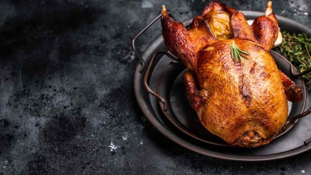 Celá pečená hus so zlatistou chrumkavou kožou na tmavom tanieri, ozdobená bylinkami.