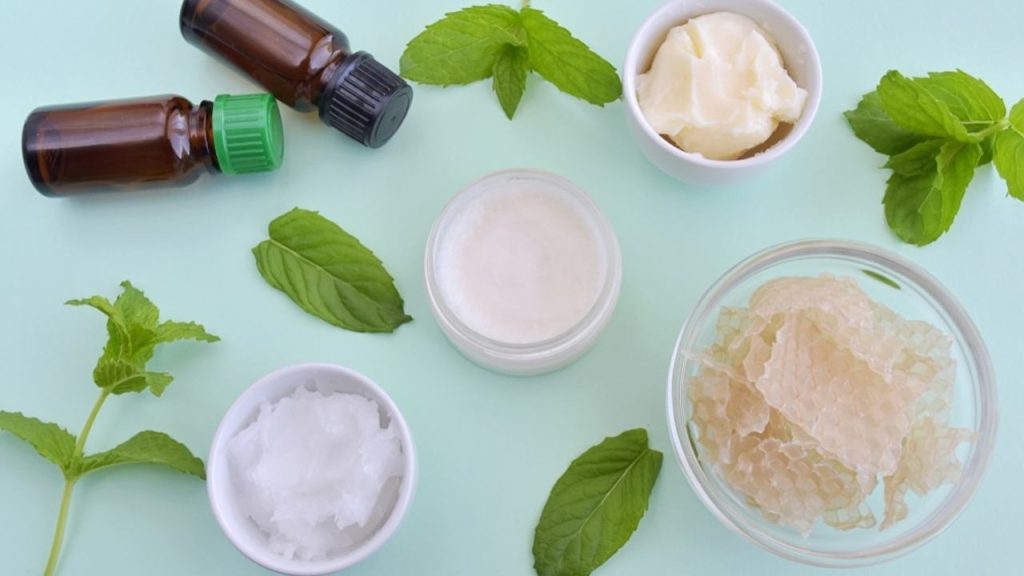 Ingrediencie na domáci balzam na pery rozložené na stole – včelí vosk, bambucké maslo, olej a bylinky.