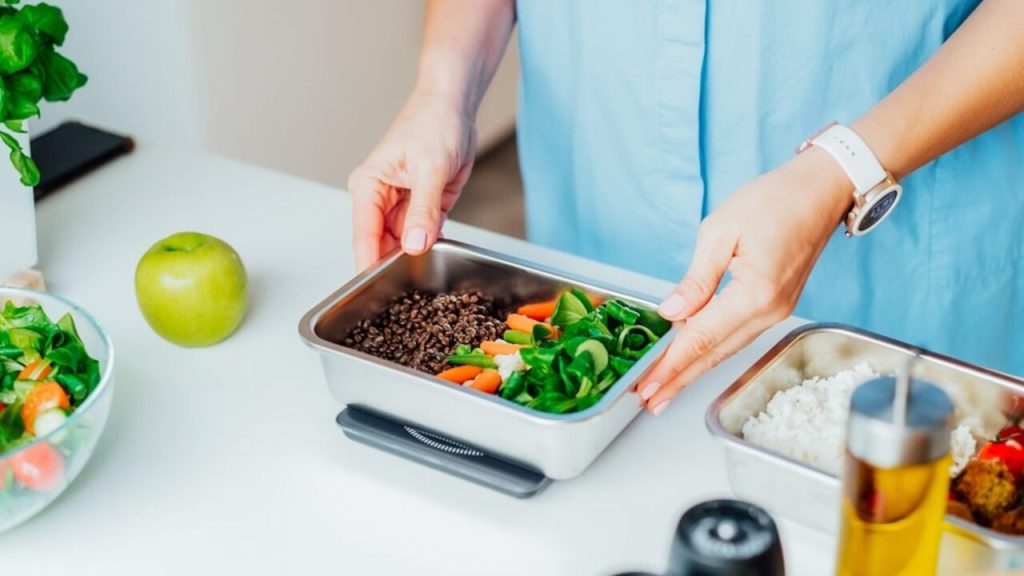Ruky ženy pripravujú krabičkový obed so zeleninou a strukovinami, položený na kuchynskej linke pri zdravých potravinách.