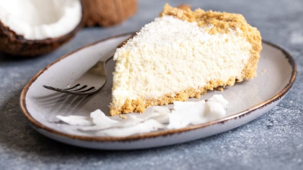 Plátok kokosového dezertu bez pečenia so sušienkovým korpusom, podávaný na tanieri s kúskami kokosu.
