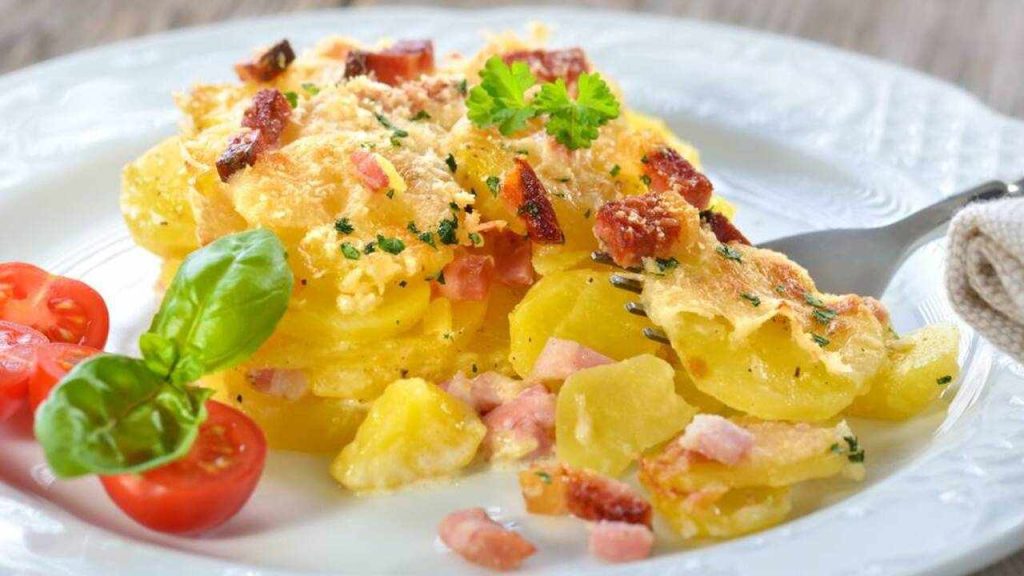 Porcia krémových zapekaných zemiakov so slaninou a syrom, podávaná na bielom tanieri s cherry paradajkami a bazalkou.