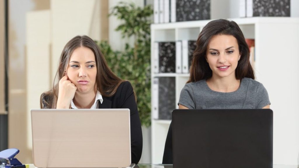 Dve kolegyne pracujú za notebookmi v kancelárii, jedna z nich sa pozerá na druhú so závistlivým výrazom.