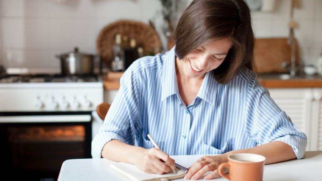 Žena sedí pri stole v kuchyni a zapisuje si poznámky do zápisníka, usmieva sa a pôsobí sústredene.