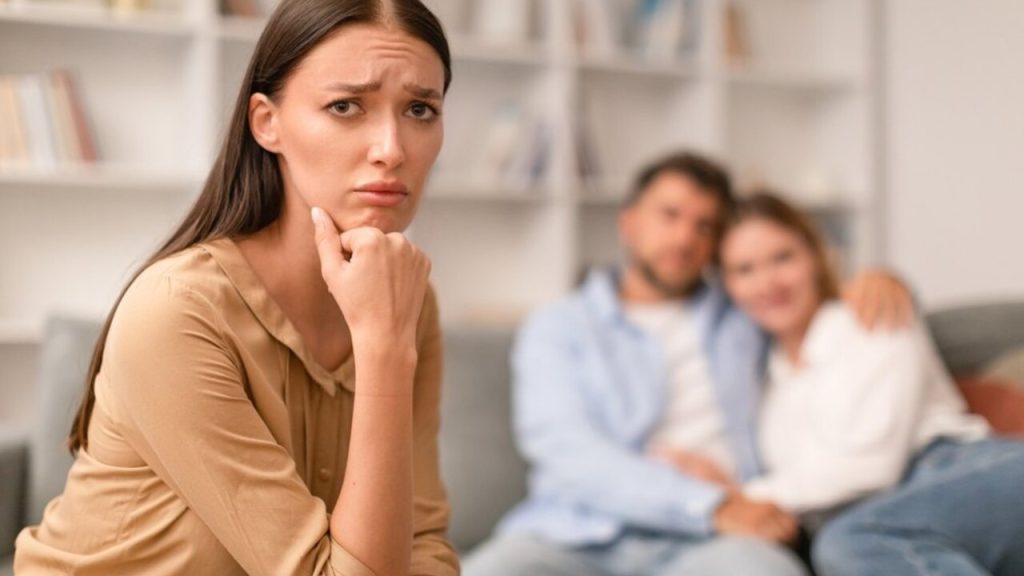 Žena so zamysleným a smutným výrazom sedí v popredí, v pozadí je šťastný pár objímajúci sa na gauči.