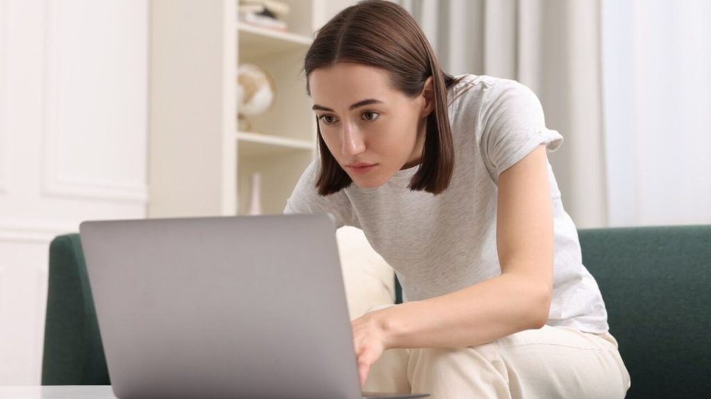 Žena zhrbená nad notebookom doma, sústredene pracuje a pôsobí unavene.