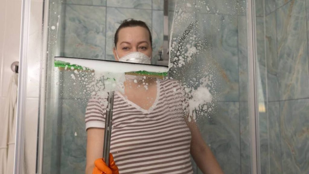 Žena čistí sklo sprchového kúta stierkou a odstraňuje penu a vodný kameň