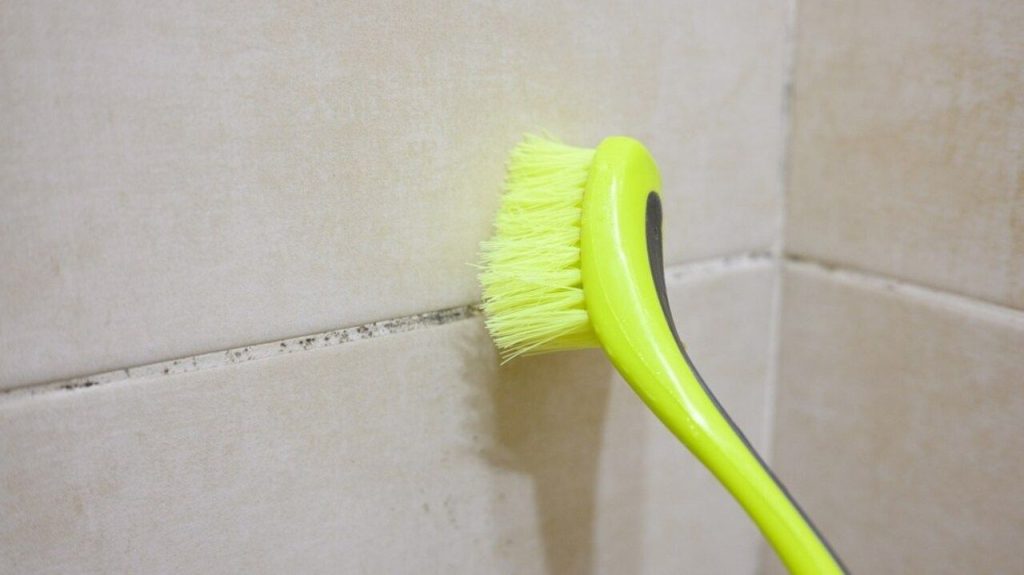Zelená kefka čistí tmavé škáry medzi béžovými kúpeľňovými dlaždicami