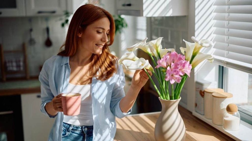 Žena pri okne aranžuje čerstvé kvety vo váze a drží šálku kávy v ruke