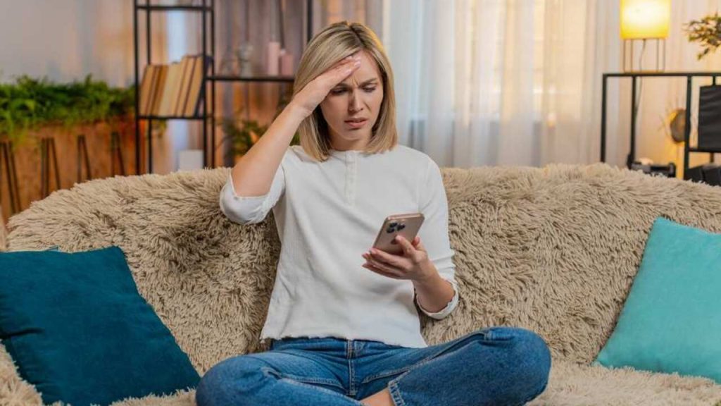 Žena sediaca na gauči doma, zamyslene sleduje mobil a drží si čelo, pôsobí vystresovane.
