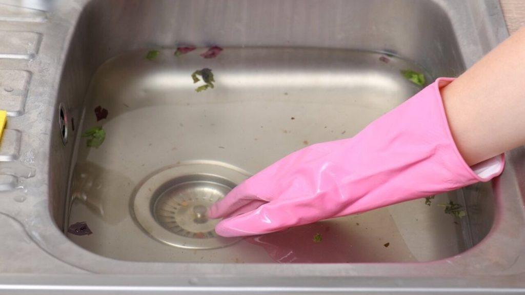 Upchatý kuchynský drez so stojacou vodou a rukou v ružovej rukavici pri odtoku