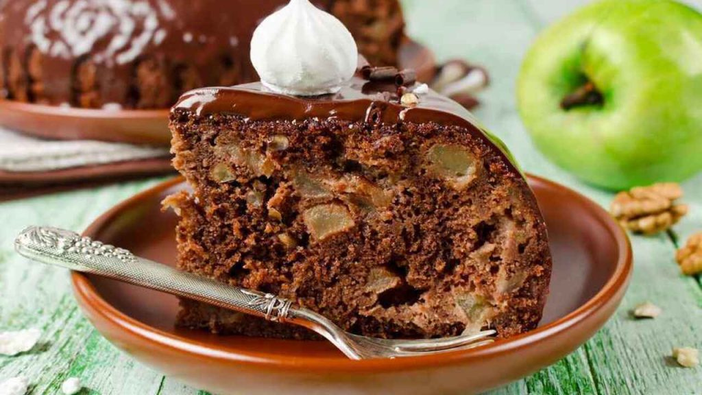 Plátok čokoládového koláča s kúskami jabĺk a orechov poliaty čokoládovou polevou na tanieri