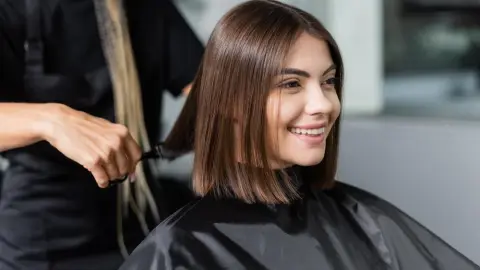 spokojnosť klienta, veselá žena s krátkymi brunetovými vlasmi sediaca v kaderníckej čiapke v salóne krásy, strihanie profesionálnym kaderníkom, salón krásy