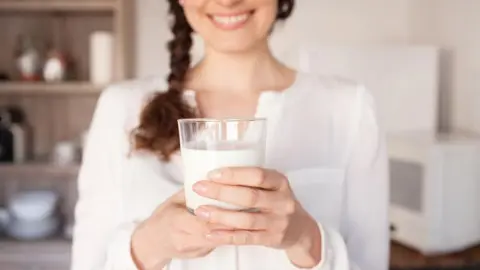 Usmievavá žena v bielej blúzke drží pohár čerstvého mlieka v kuchyni.