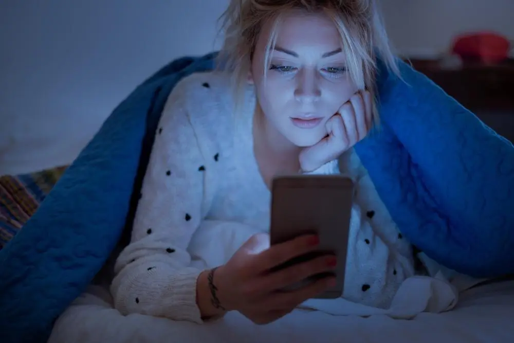 Mladá žena v posteli pozerajúca do mobilu neskoro v noci, ilustrujúca škodlivý návyk nedostatku spánku.