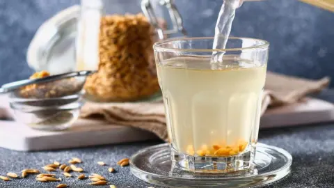 Pohár ovseného nápoja, do ktorého sa leje voda, na stole sú ovsené zrná a sitko