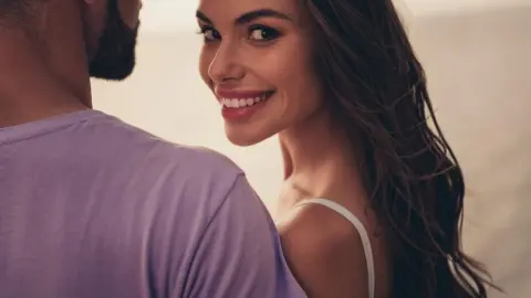 Usmiata žena objímajúca partnera, pohľad do kamery, romantický moment pri západe slnka