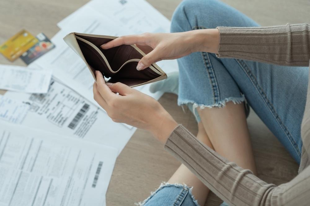 Žena sedí na podlahe s prázdnou peňaženkou v rukách, okolo nej účty a kreditné karty – symbol finančných problémov.