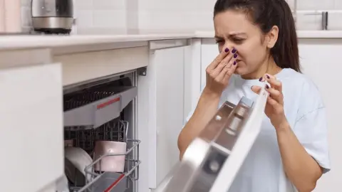 Žena drží otvorené dvierka umývačky riadu a zakrýva si nos pred nepríjemným zápachom