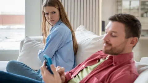 Žena sedí vedľa partnera, ktorý si ju nevšíma a venuje sa mobilu, zatiaľ čo ona ho pozoruje s pocitom frustrácie