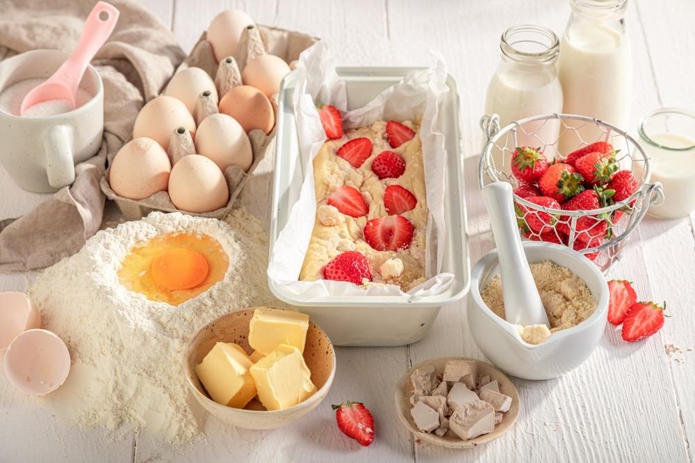 Ingrediencie na domáci jahodový koláč – múka, vajcia, maslo, cukor, mlieko, droždie a čerstvé jahody pripravené na pečenie.