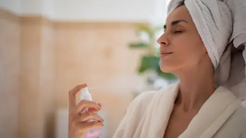 Žena v župane si strieka na tvár osviežujúci sprej ako súčasť letnej starostlivosti o pleť.