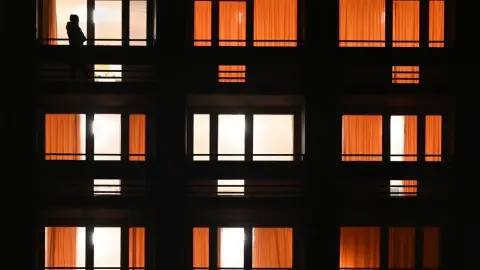 Silueta človeka na balkóne pred rozsvietenými oknami bytového domu večer.