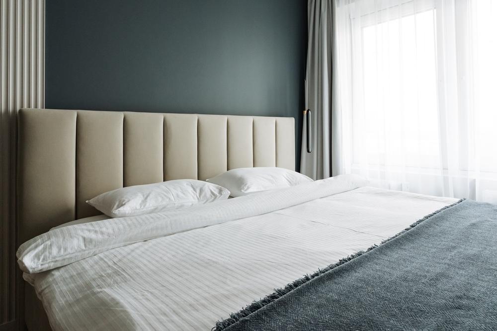 Moderná spálňa so sivomodrou stenou, béžovým čelom postele a bielou posteľnou bielizňou, zaplavená denným svetlom.