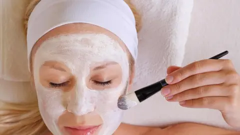 Žena s bielou pleťovou maskou na tvári si nanáša masku štetcom počas domácej starostlivosti o pleť