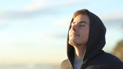 Mladý muž v mikine si vychutnáva ranné slnko so zatvorenými očami.