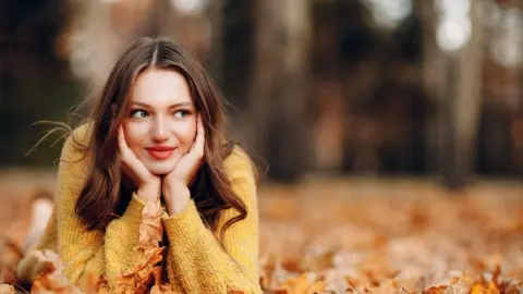 Usmievavá mladá žena v jesennom parku, zamyslene leží na lístí a pozerá do diaľky.