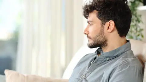 Mladý muž sedí sám na gauči a zamyslene hľadí do diaľky. Symbol osamelosti a izolácie.