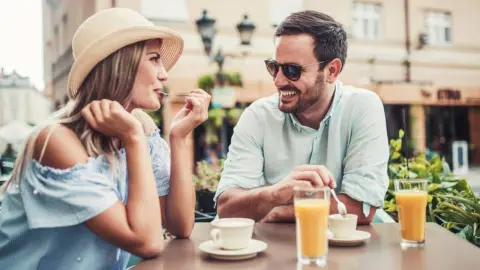 Muž a žena sa rozprávajú na terase kaviarne, obaja sa usmievajú – medzi nimi cítiť flirt a iskru.