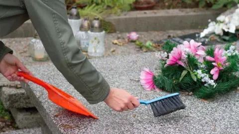 Človek čistí hrob metličkou a lopatkou, na kameni ležia umelé kvety.