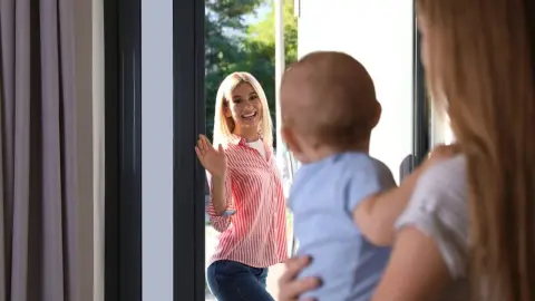 Mama sa lúči s dieťaťom pri dverách, usmieva sa a máva – pokojný a láskavý moment plný dôvery.
