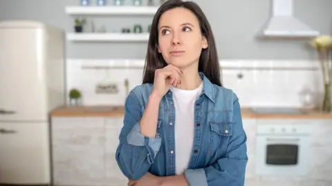 Zamyšlená žena stojí v kuchyni a premýšľa, prečo tam prišla.