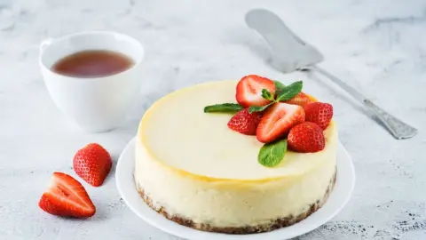 Krémový cheesecake s jahodami a mätou na bielom tanieri, vedľa šálky čaju.