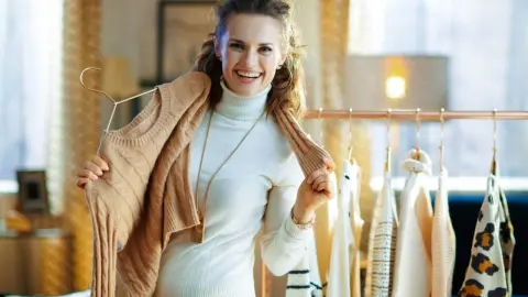 Usmiata žena drží béžový sveter a vyberá si oblečenie zo svojho jesenného šatníka