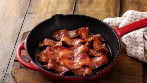 Chrumkavé plátky slaniny v liatinovej panvici na drevenom stole.