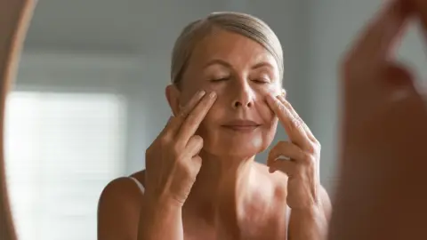Žena si pred zrkadlom robí lymfatickú masáž tváre ako súčasť rannej rutiny.