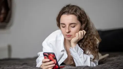 Mladá žena leží na posteli a pozerá do mobilného telefónu.