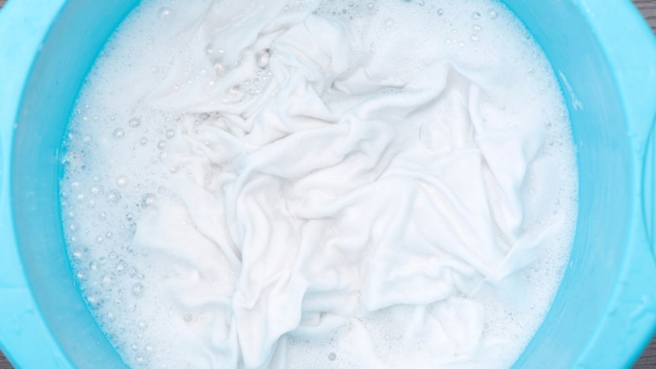 Biela bielizeň namočená v mydlovej pene v modrom vedre počas čistenia.