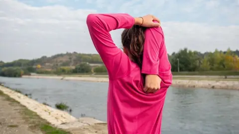 Žena v ružovej športovej mikine si naťahuje ruku za hlavou počas strečingu pri rieke.